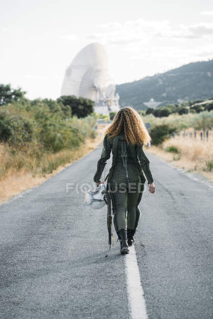 Rückansicht einer Frau im Astronautenkostüm, die die Landstraße entlanggeht — Stockfoto