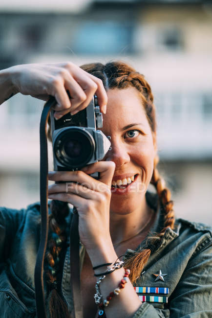 Retrato de chica pelirroja sonriente con trenzas tomando fotos con cámara vintage - foto de stock