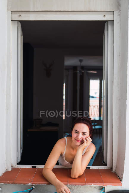 Jovem sorrindo mulher de pé na janela e olhando para a câmera — Fotografia de Stock