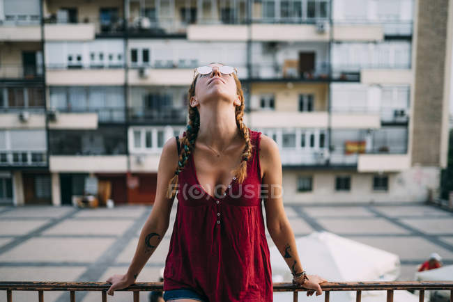 Linda chica pelirroja con trenzas y gafas de sol apoyadas en barandilla contra edificio residencial - foto de stock