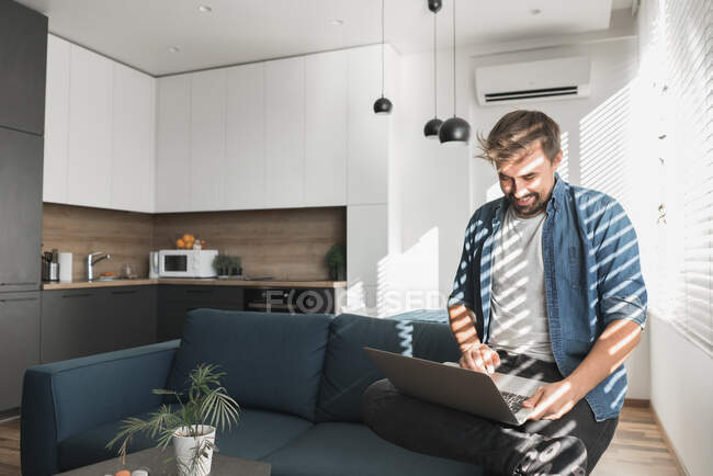 Bearded guy browsing modern laptop while sitting on comfortable couch in stylish living room — Stock Photo