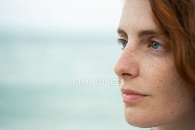 Schöne junge Frau mit Ingwerhaaren schaut weg, während sie auf verschwommenem Hintergrund von Strand und Meer in Tyulenovo, Bulgarien steht — Stockfoto