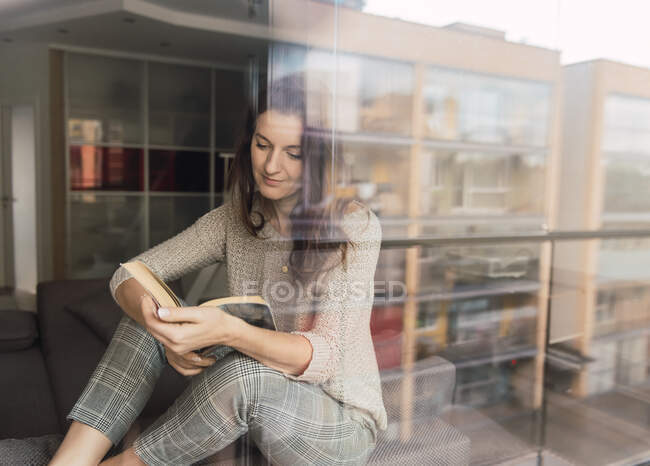 Красивая взрослая женщина в элегантном наряде улыбается и смотрит в сторону, лежа на удобном диване с интересной книгой — стоковое фото