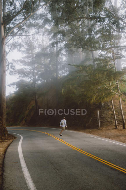Vue arrière d'un jeune homme anonyme chevauchant une planche à roulettes le long d'une route asphaltée dans une magnifique forêt lors d'une journée brumeuse à Big Sur, Californie — Photo de stock