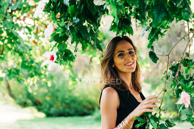 Mujer alegre y elegante de pie bajo el árbol y sonriendo a la cámara en el parque - foto de stock