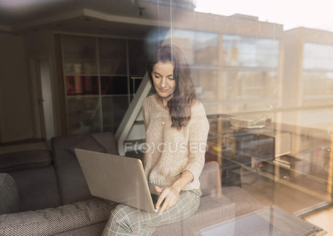 Красивая взрослая женщина в элегантном наряде просматривает современный ноутбук и смотрит в камеру, сидя в уютной комнате за огромным оконным стеклом — стоковое фото