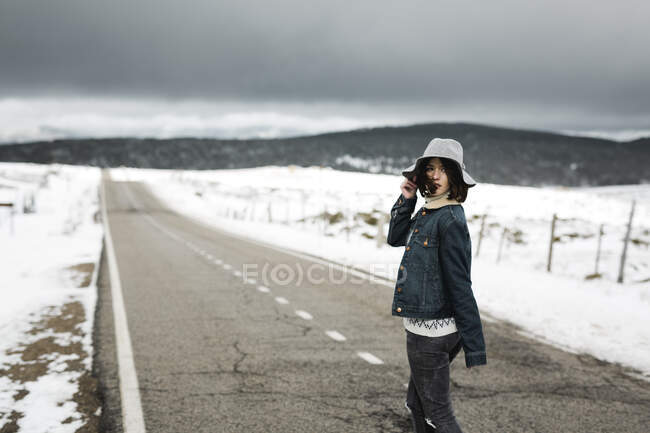 Rückansicht eines jungen Menschen in stylischem Outfit, der an bewölkten Wintertagen mitten auf einer asphaltierten Straße in einer wunderschönen Landschaft spaziert — Stockfoto