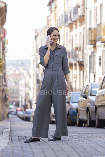 Красивая женщина в элегантном наряде улыбается и отворачивается, стоя на городской улице и разговаривая на смартфоне — стоковое фото
