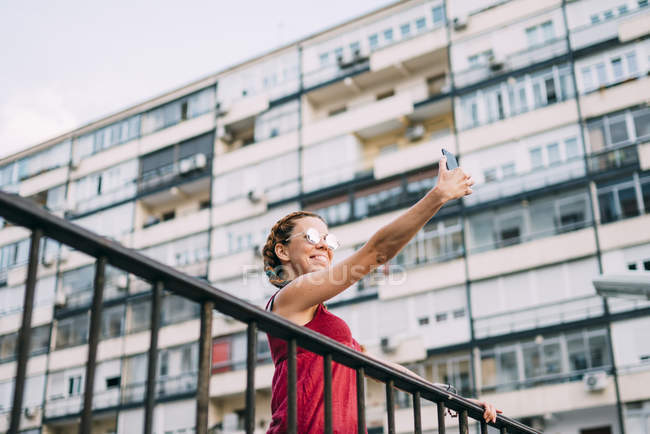 Smiling red-haired girl with braids taking photo with mobile phone against residential building — Stock Photo