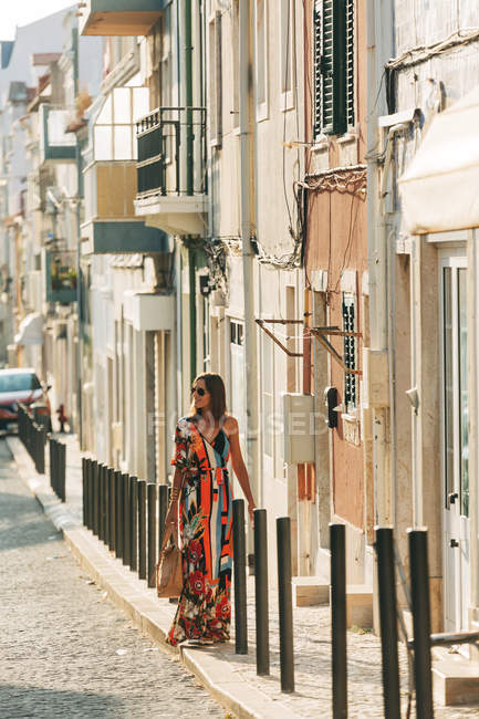 Elegant woman in long dress carrying handbag and walking on street in sunlight — Stock Photo