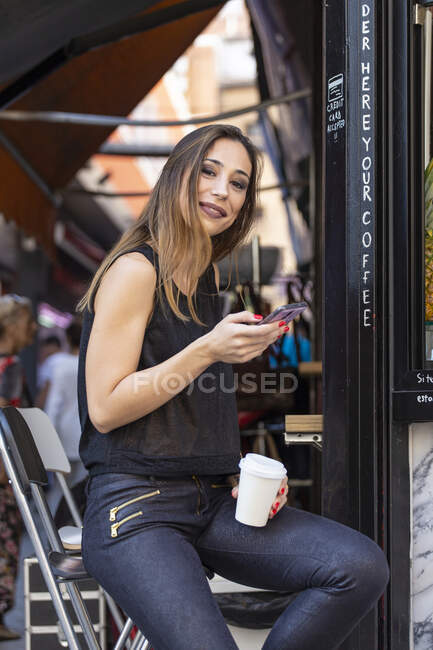 Приваблива жінка з чашкою гарячого напою і сучасним смартфоном посміхається і дивиться на камеру, сидячи біля кафе — стокове фото
