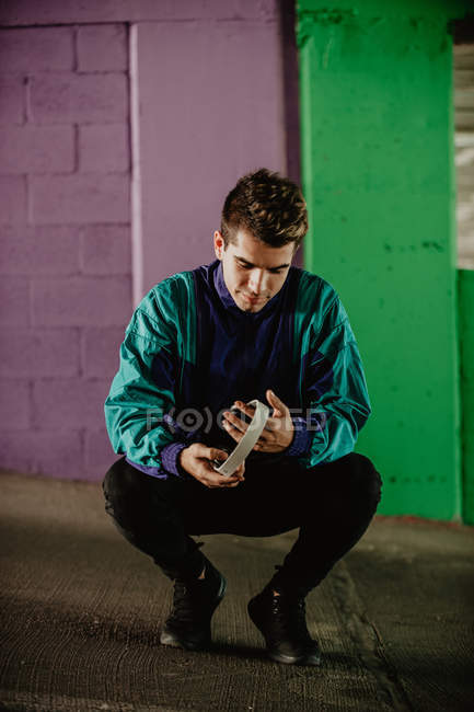 Jeune homme en vêtements de sport avec écouteurs accroupi contre un mur coloré — Photo de stock
