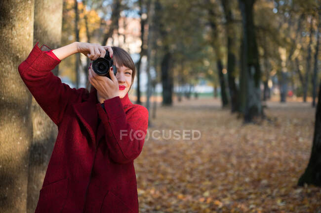 Молода леді в червоному пальто знімає на цифровій камері в осінньому лісі — стокове фото