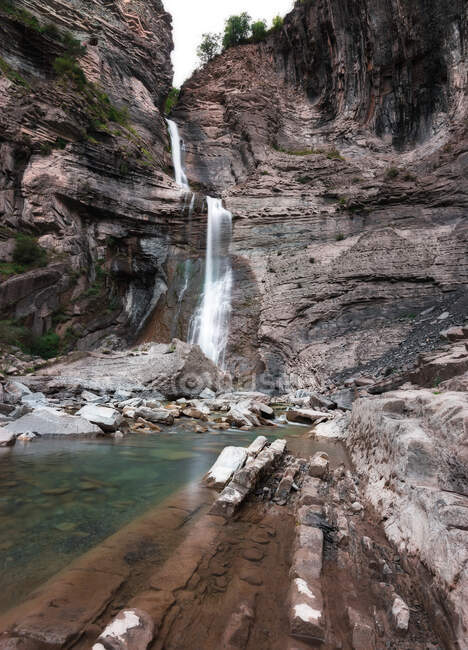 Висока крута скеля з маленьким каскадним водоспадом, що тече в чисту річку — стокове фото