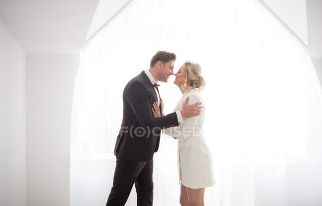Young handsome man in black costume and beautiful woman in white jacket standing in room near window and hugging — Stock Photo