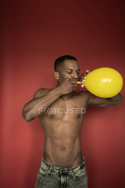 Мускулистый черный мужчина в джинсах, дующий ярко-желтым шариком на красном фоне — стоковое фото