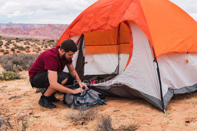 Vista laterale di bel ragazzo in abito casual imballaggio zaino mentre seduto a terra vicino tenda sulla zona campeggio nel deserto — Foto stock