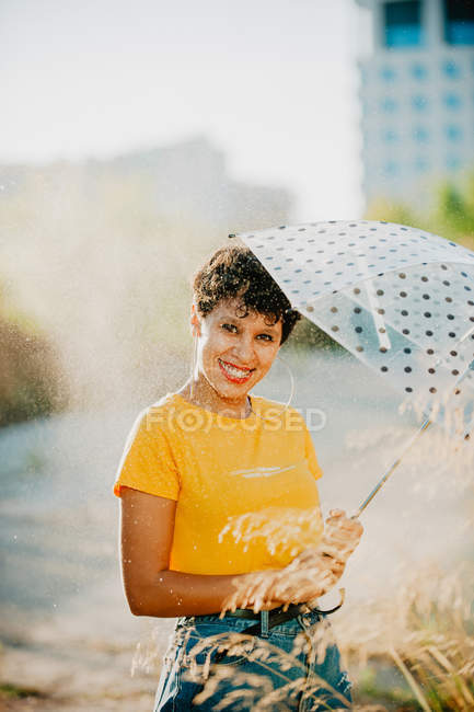 Portrait of smiling woman in stylish outfit with umbrella standing under drops of spraying water — Stock Photo