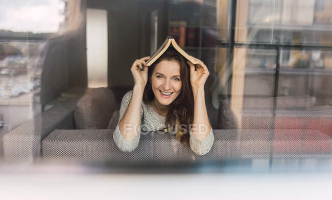 Lovely adult lady in elegant outfit smiling and looking at camera while lying on comfortable sofa with interesting book — Stock Photo