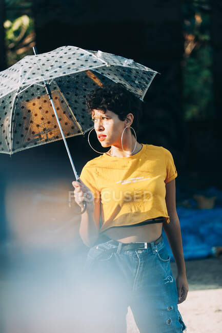 Lovely young woman in stylish outfit holding umbrella and looking away while standing on street during rain on sunny day — Stock Photo