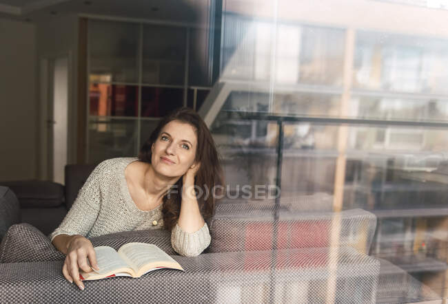 Schöne erwachsene Dame in elegantem Outfit lächelt und schaut weg, während sie auf einem bequemen Sofa mit interessantem Buch liegt — Stockfoto