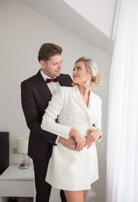 Joven hombre guapo en traje negro y hermosa mujer en chaqueta blanca de pie en la habitación cerca de la ventana y abrazos - foto de stock
