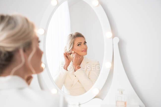 Back view of young beautiful woman in white formal costume with hairdo standing in room in front of dressing table and looking in mirror — Stock Photo