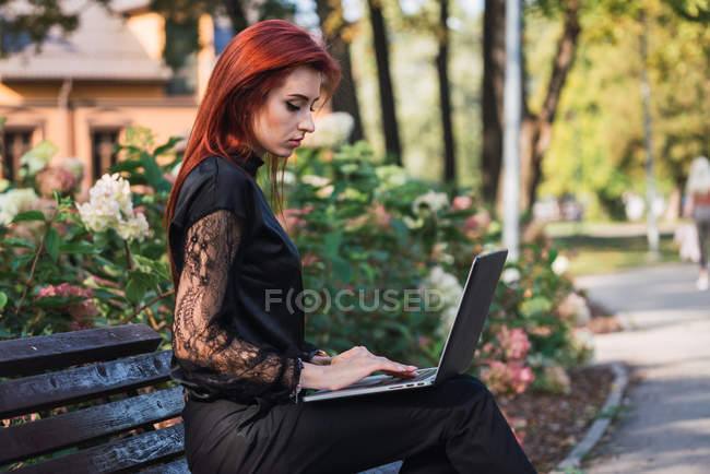 Mujer joven elegante usando el ordenador portátil en el banco en el parque - foto de stock
