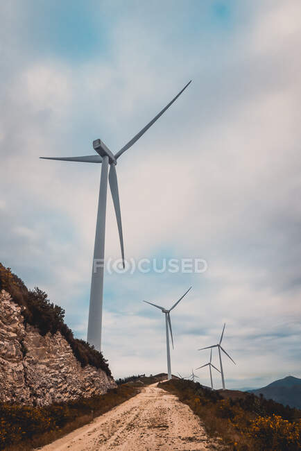 Fila de moinhos de vento modernos de pé no lado da estrada rural estreita no dia nublado — Fotografia de Stock
