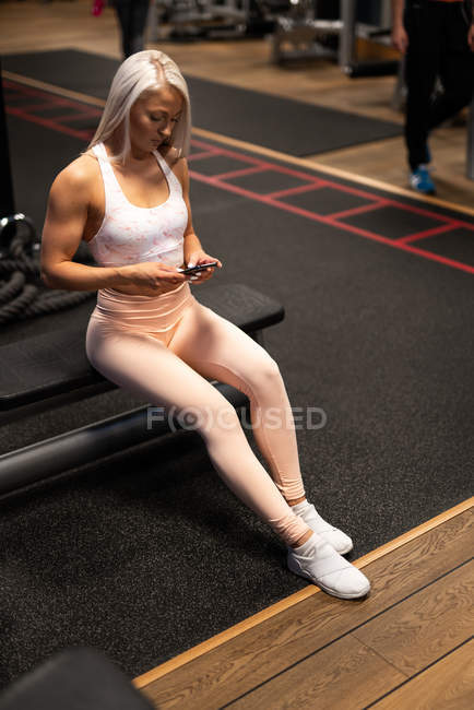 Blond athletic woman in sportswear and sneakers sitting in gym and using smartphone — Stock Photo