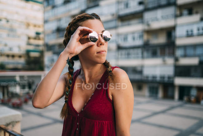 Стильная рыжая девушка с косичками и солнцезащитными очками стоит в городе — стоковое фото