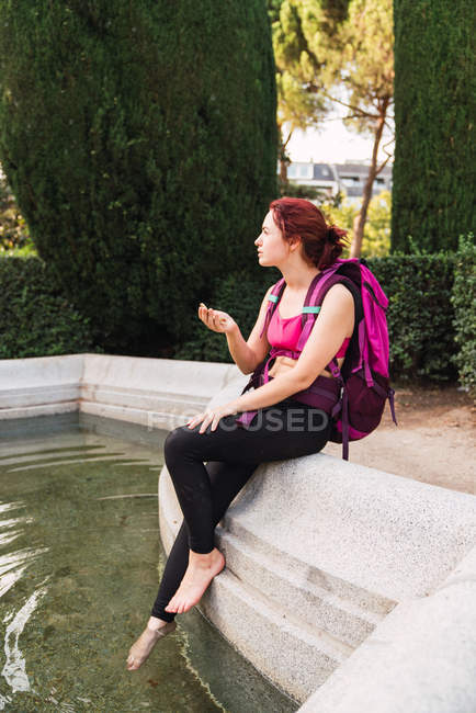 Young woman with backpack sitting on parapet near lake with compass in hand — Stock Photo