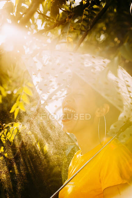 Side view of attractive young female with umbrella looking at camera while standing under wet tree branches on sunny day in jungle — Stock Photo