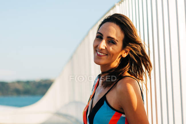 Jolie femme brune appuyée sur la clôture du pont et regardant la caméra en plein soleil — Photo de stock