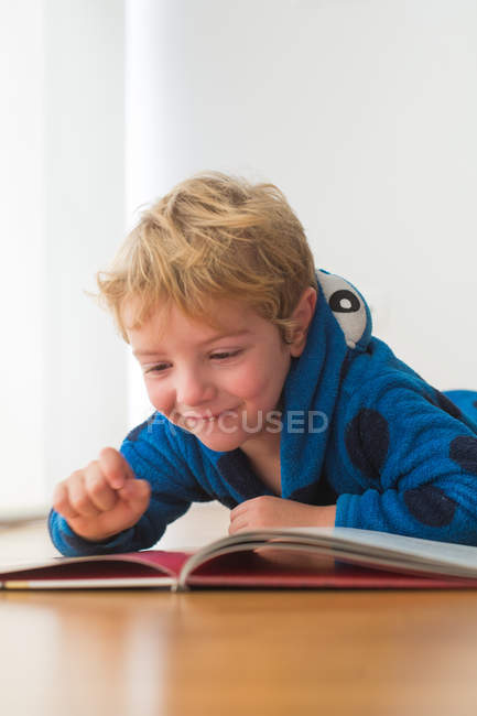 Smiling blonde little boy reading book on wooden floor — Stock Photo