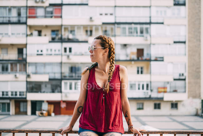 Menina ruiva bonita com tranças e óculos de sol apoiados em trilhos contra edifício residencial — Fotografia de Stock
