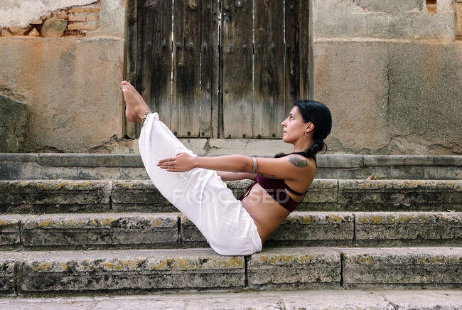 Barefoot woman sitting in Boat pose while doing yoga on weathered steps of ancient building — Stock Photo