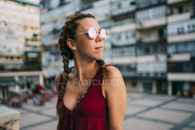 Стильная рыжая девушка с косичками и солнцезащитными очками стоит в городе — стоковое фото