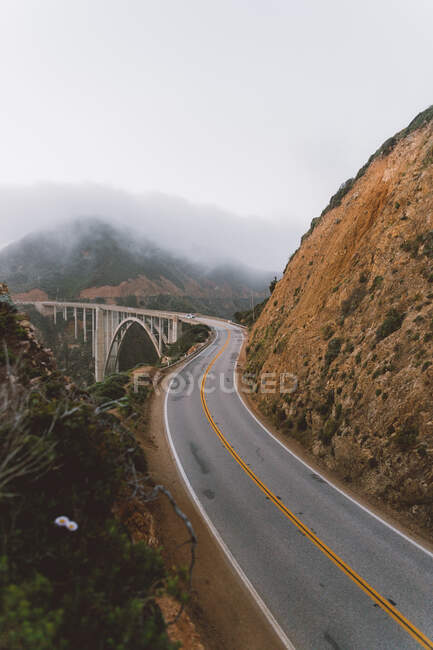 Carretera estrecha de asfalto y hermoso puente ubicado cerca de la ladera de la montaña en el día de niebla en Big Sur, California - foto de stock