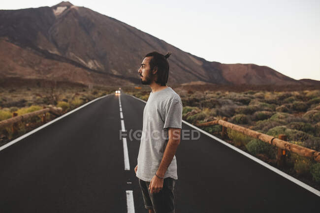 Vista lateral de homem barbudo bonito em roupa casual em pé na estrada de asfalto passando pela magnífica ilha de Tenerife, na Espanha — Fotografia de Stock