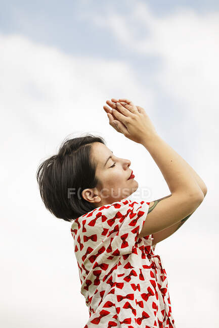 From below view of attractive brunette woman in patterned shirt with tattoos on arm standing on background of cloudy sky with closed eyes — Stock Photo
