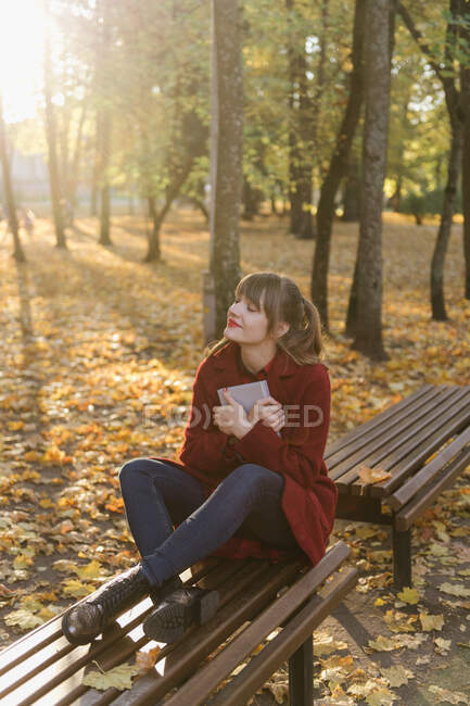 Jovem atraente de casaco vermelho segurando livro aberto e sentado no assento na floresta de outono — Fotografia de Stock