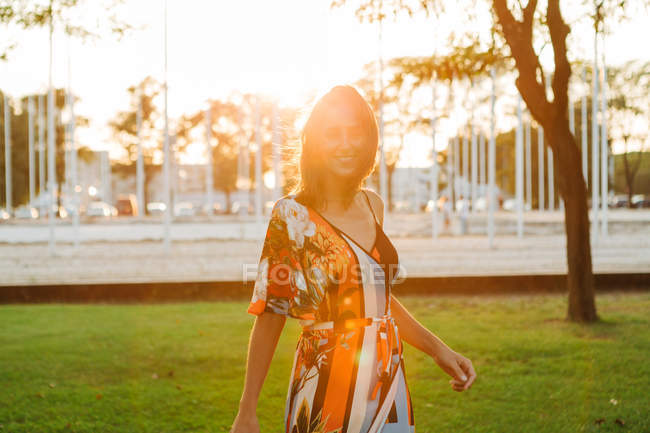 Веселая стильная брюнетка в платье, стоящая на зеленой лужайке в городском парке и улыбающаяся в камеру — стоковое фото