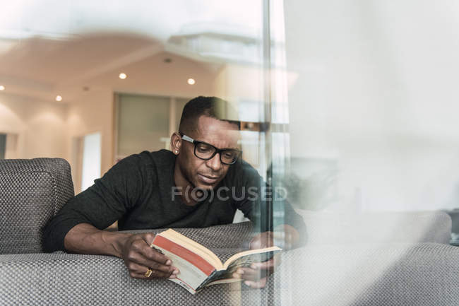 Цілеспрямований афроамериканець людиною, в окулярах, читання книги відпочиваючи на диван в домашніх умовах — стокове фото
