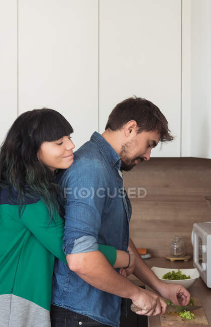 Вид збоку красива молода жінка тримає очі закритими і обіймає кулінарного чоловіка ззаду, стоячи в стильній кухні разом — стокове фото