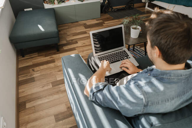 Зверху знімок молодого хлопця, який переглядає сучасний ноутбук з чорним екраном, сидячи на зручному дивані у стильній вітальні — стокове фото