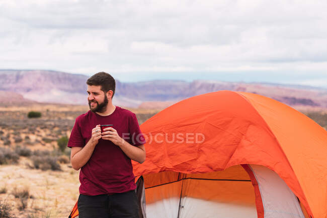 Bearded guy in casual outfit holding mug of hot beverage and modern smartphone while sitting near tent and looking away in beautiful nature — Stock Photo