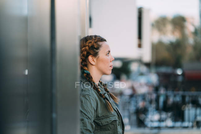 Thoughtful red-haired girl with braids leaning on wall in city — Stock Photo