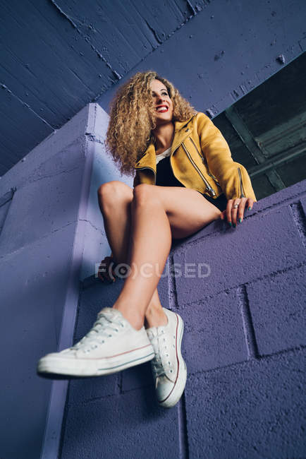 Стильная кудрявая блондинка в кроссовках и желтой куртке сидит на фиолетовой стене и смеется — стоковое фото