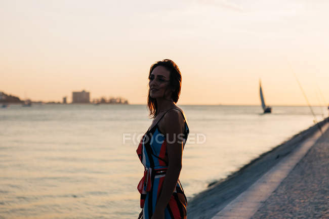Mujer joven soñadora en vestido de pie en el paseo marítimo adoquinado al atardecer contra el paisaje marino y mirando a la vista - foto de stock